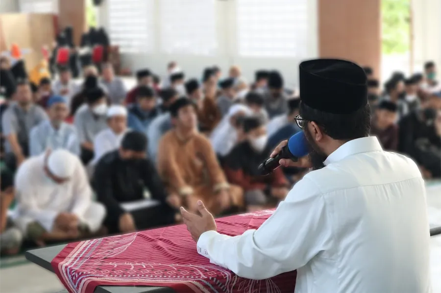 Ilustrasi mimpi bertemu ulama — a person in a white shirt and black hat speaking into a microphone