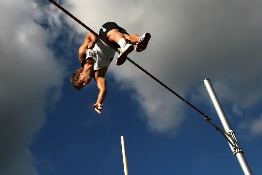 Ilustrasi mimpi loncat galah — man in white tank top and black shorts hanging on black metal bar under blue sky