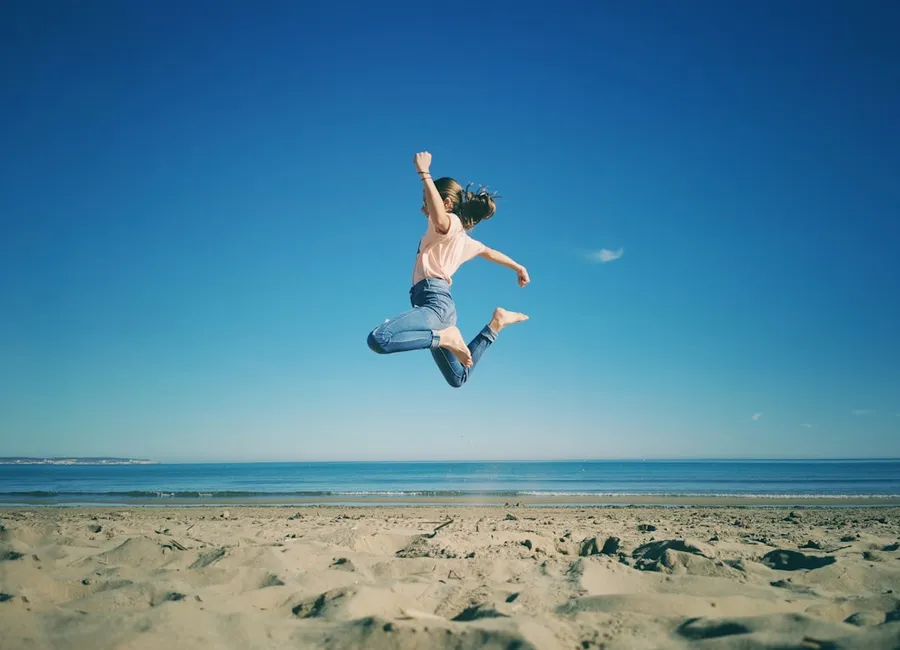 Ilustrasi mimpi loncat tinggi — woman in white tank top and blue denim shorts jumping on beach during daytime