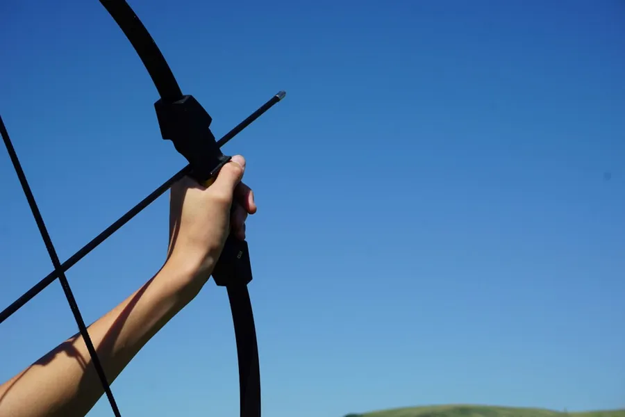 Ilustrasi mimpi panahan — a hand holding a bow and arrow in front of a blue sky