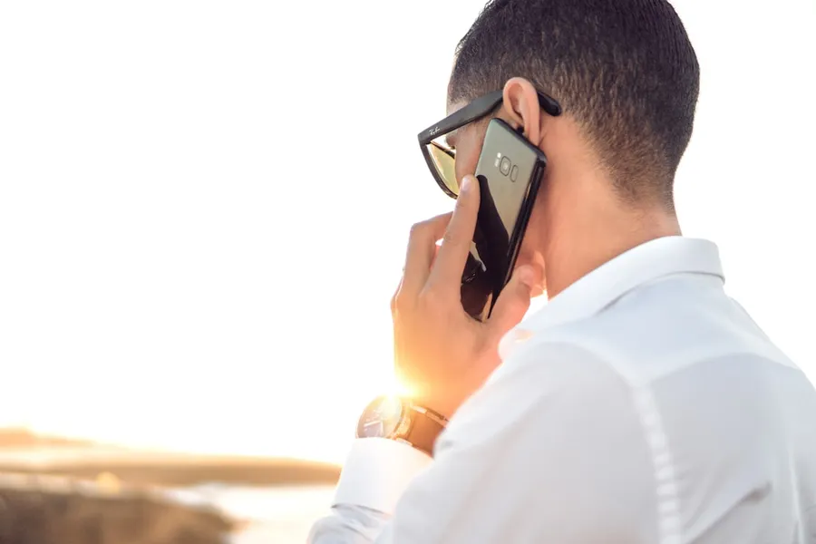 Ilustrasi mimpi menelpon — man holding smartphone standing in front of calm body of water