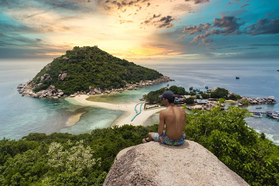 Ilustrasi mimpi orang thailand — topless man sitting on cliff rock viewing Ko Nang Yuan isalnd, houses, and buildings under white and yellow sky
