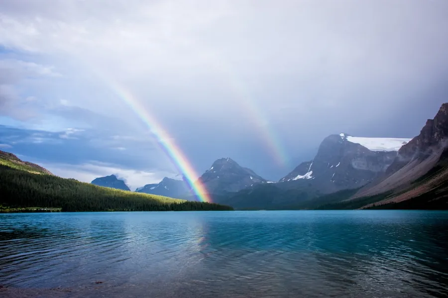 Ilustrasi mimpi pelangi ganda — blue lake and rainbow under nimbus clouds