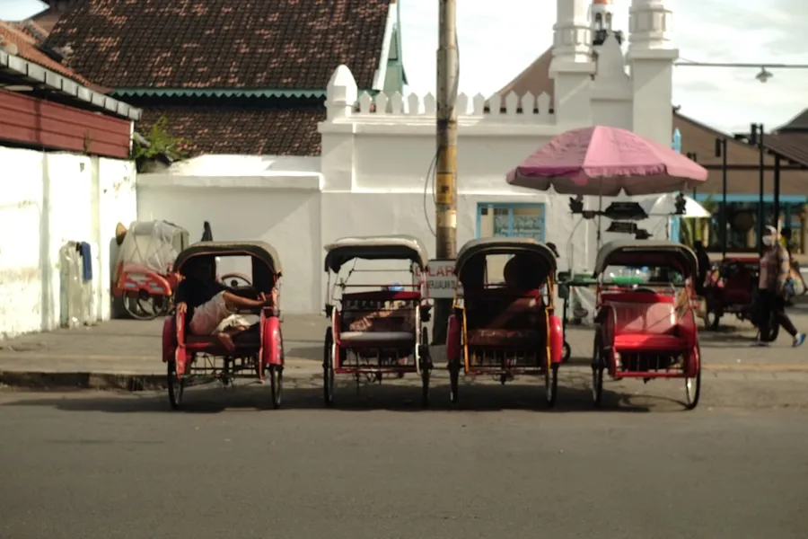 Ilustrasi mimpi tukang becak — Rickshaws line a street near a white building.
